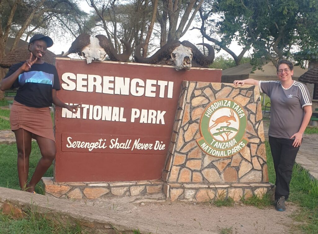 People standing either side of the Serengeti National Park entrance sign.